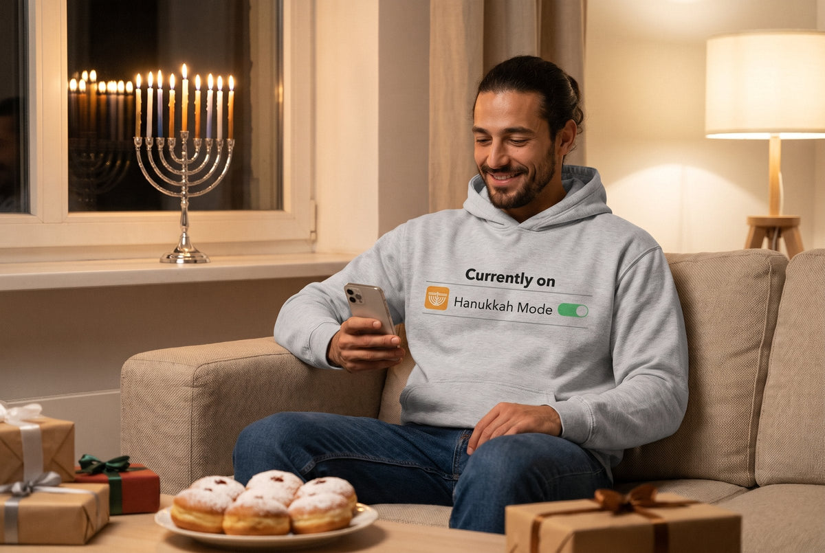 Hanukkah Mode hoodie worn by a man smiling at his phone on a sofa next to a lit Hanukkah menorah and sufganiyot – MoT Member of the Tribe