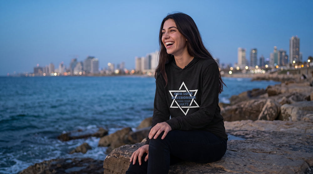 Our Love Is Stronger hooded long sleeve shirt worn by a woman on the Jaffa shore at blue hour – MoT Member of the Tribe